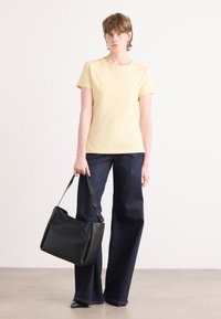 T-shirt di cotone gialla a maniche corte, abbinata a jeans svasati blu scuro. Una borsa a spalla in pelle nera con tracolla larga è tenuta in mano.