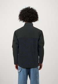 Giacca nera e texturizzata con motivi a onde blu sul retro, colletto alto e maniche lunghe; indossata con pantaloni in denim larghi.