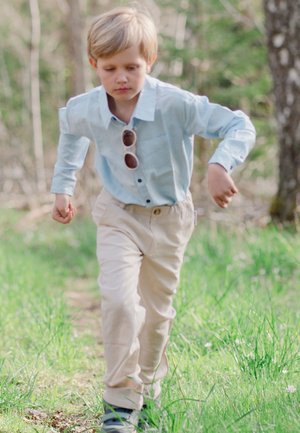 Lysblå skjorte med knapper, beige bukser og solbriller; barn løber på en græsklædt sti i en naturlig omgivelser med blødt sløret baggrund.