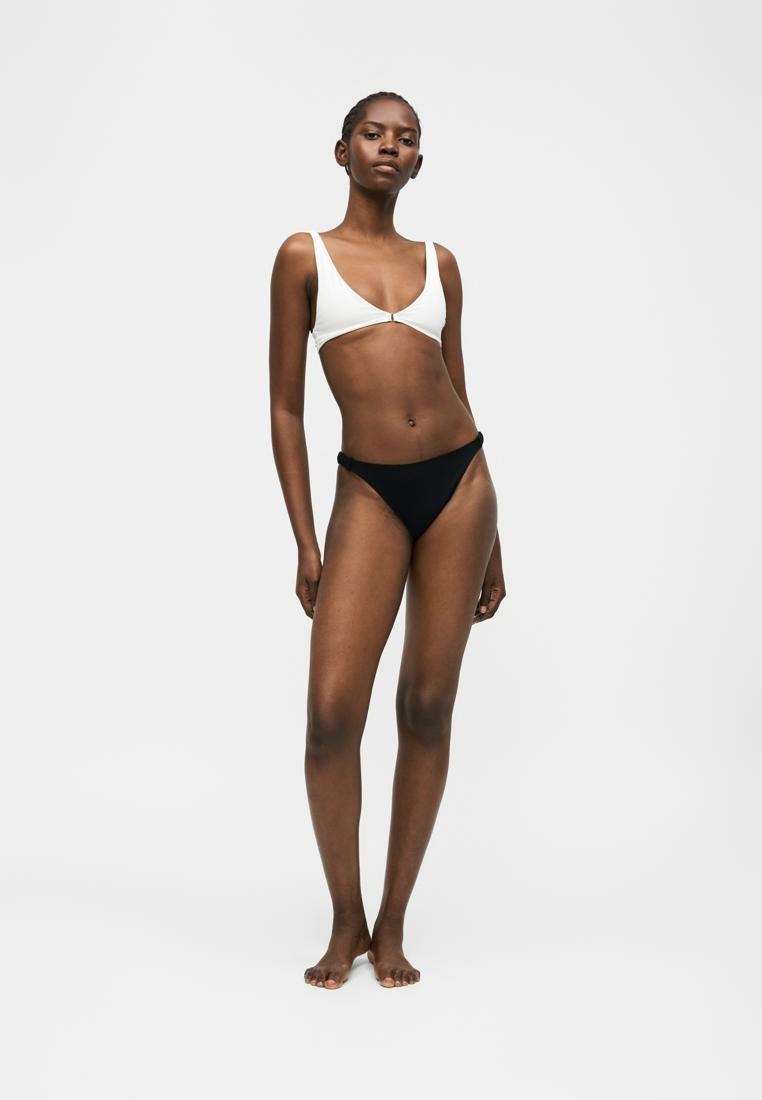 Femme debout pieds nus portant un haut de bikini blanc et un bas de bikini noir sur un fond clair uni.