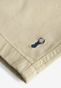 Tissu beige avec des coutures détaillées et une petite tête de cerf brodée en bleu marine près du bord.