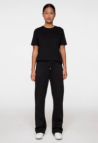 Musta puuvillainen T-paita, jossa on rento istuvuus, yhdistetty mustiin nyörillisiin collegehousuihin. Mallilla on valkoiset tennarit. Neutraali tausta.