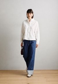 Blouse blanche à manches longues avec un motif texturé et ondulé, associée à un jean en denim bleu ample. Modèle tenant un petit sac marron et portant des baskets.