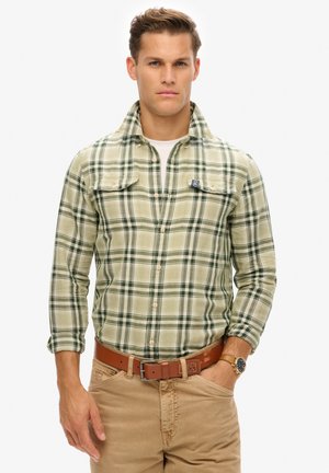 Chemise à carreaux verte et crème avec col, deux poches avant et fermeture à boutons, portée par-dessus un t-shirt blanc. Ceinture marron et montre au poignet visibles.