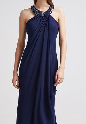 Woman wearing a sleeveless navy blue gown with a beaded halter neckline and flowing fabric draped at the front.
