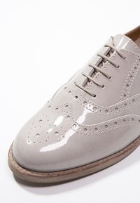 Chaussure brogue à lacets en cuir verni gris clair avec détails perforés, bout arrondi et semelle marron contrastante.