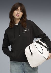 Sort hættetrøje med emblem, der viser "PUMA ATHLETICS", og grå jeans. Holder en hvid taske med sorte detaljer og logo. Glat stof, afslappet design.