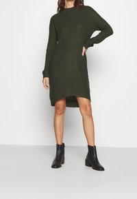 Abito in maglia di colore verde scuro, caratterizzato da un collo alto, maniche lunghe e un orlo asimmetrico. Abbinalo a stivali neri alla caviglia. Tessuto con texture.