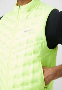 Gilet imbottito senza maniche di un brillante verde lime con logo Nike argentato, dotato di tasche con zip e una texture liscia e leggera.