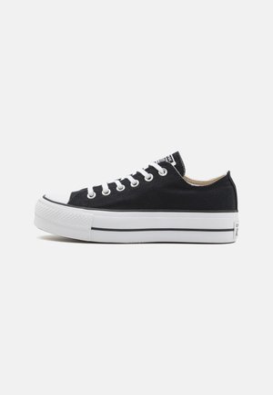 Zapatillas bajas de lona negra con punta de goma blanca, cordones y suela. Presentan una suela texturizada y una franja negra. Diseño clásico.