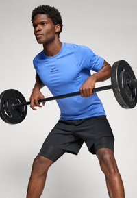 Un homme portant un t-shirt bleu Nike Pro et un short noir soulève une barre pondérée, se préparant pour un exercice de tirage penché.