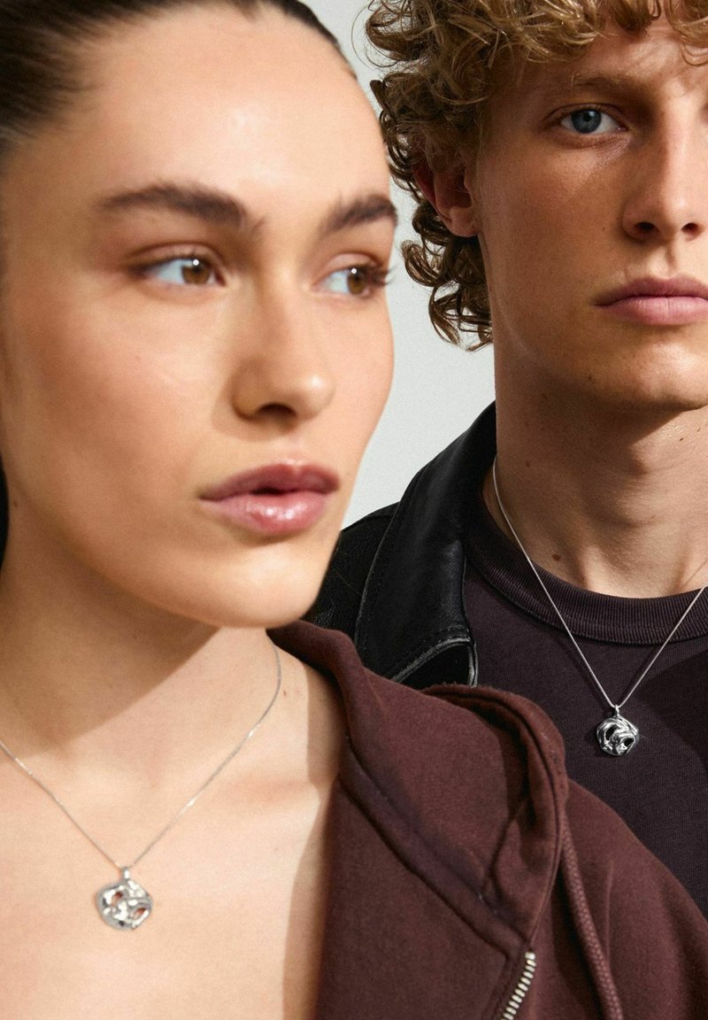 Silverhalsband med strukturerade hängen på personer som bär en burgundy huvtröja och en mörk jacka, mot en neutral bakgrund.