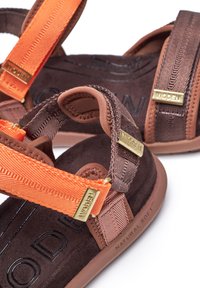 Brown textile sandals with orange straps. Features gold hardware accents and textured surfaces. Soft inner material with printed logo.