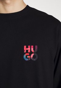 Schwarzer Rundhalsausschnitt-Pullover mit buntem "HUGO"-Textlogo auf der Brust, das Rot-, Lila- und Blautöne kombiniert.