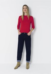 Rotes Langarmshirt, dunkelblaue Jeans mit umgeschlagenen Säumen und beigen Knöchelboots. Einfaches Design, glatte Textur und gerader Schnitt bei den Jeans.