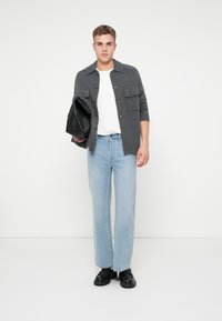 Grauer Cardigan mit Knöpfen und zwei Taschen, weißes T-Shirt, hellblaue Jeans und schwarze Schuhe, hält eine schwarze Tasche. Einfaches, lässiges Outfit.