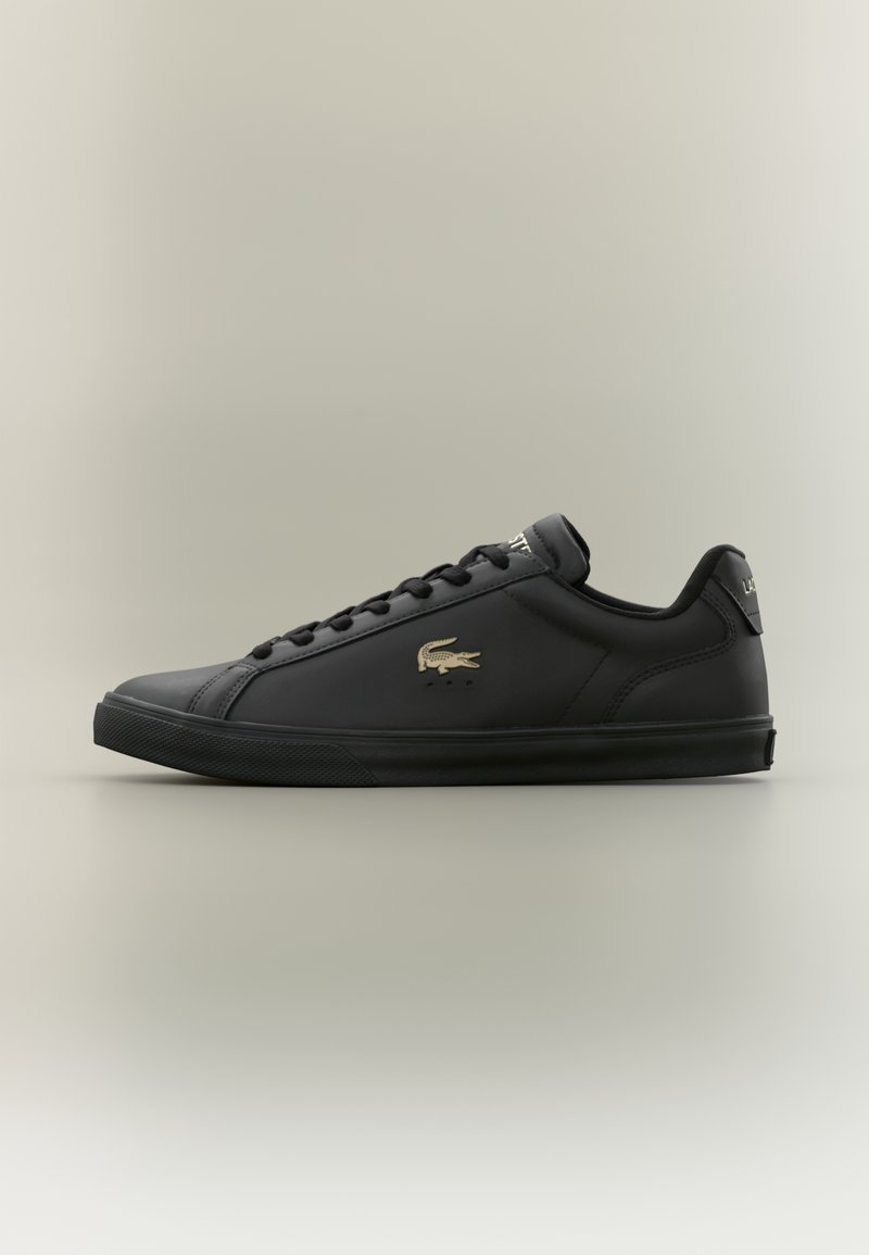 Baskets basses noires avec des lacets noirs, une semelle texturée et un logo crocodile doré sur le côté, présentées sur un fond clair.