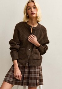 Brązowy dziergany kardigan z okrągłym dekoltem, dwiema przednimi kieszeniami i zapięciem na guziki, połączony z plaidową spódnicą w odcieniach brązu i kremu.