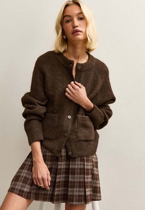 Cárdigan de punto marrón con cuello redondo, dos bolsillos frontales y cierre de botones, combinado con una falda a cuadros en tonos marrones y crema.