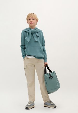 Haut à capuche en tricot bleu clair, pantalon cargo beige, baskets grises. Tient un sac doux en teal avec des anses noires. Design simple, look décontracté.