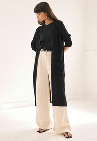 Lång svart cardigan med öppen framsida, ribbade ärmslut och sidofickor, ihopparad med beiga vida byxor och svarta flip-flops.