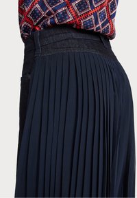 Pantalons plissés navy avec une ceinture accentuée en denim foncé. Le tissu a une texture lisse, avec des plis verticaux distincts.