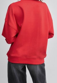 Červená mikina s volným střihem, vysokým límcem, spadlými rameny a žebrovanými manžetami. Hladká textura, minimální detaily.