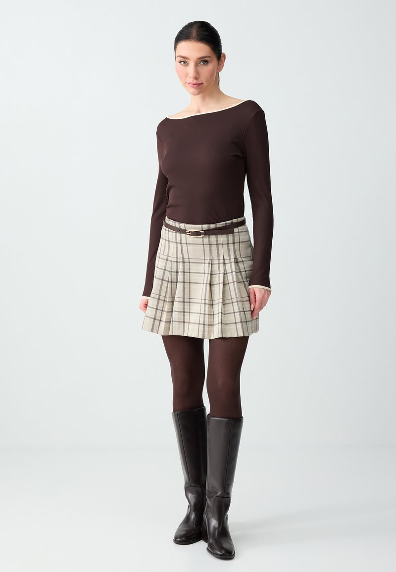 Femme portant un haut à manches longues marron, une jupe plissée beige à carreaux avec ceinture, des collants marron et des bottes noires montantes, debout devant un fond clair.