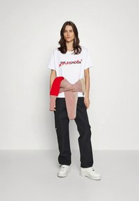 Valkoinen puuvillainen t-paita, jossa graafista tekstiä, yhdistetty mustiin cargo-housuihin. Vyötärölle sidottu monivärinen neulottu neule tuo tekstuuria. Valkoiset paksupohjaiset tennarit.