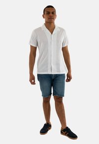 Camisa de manga corta blanca y con botones, hecha de una tela ligera, combinada con pantalones cortos de mezclilla azul y zapatillas negras con detalles en marrón.