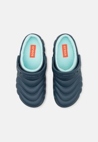 Des Crocs bleu marine avec un motif de vagues texturé, dotés d'un intérieur doux vert menthe et d'une étiquette de marque rouge sur la semelle intérieure.