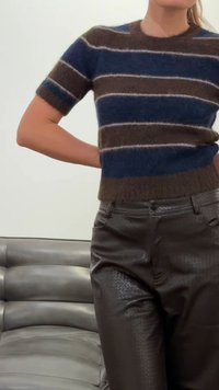 Kvinne iført en blå og brun stripete, plysj kortermet genser og teksturerte mørkebrune skinnbukser, stående ved et grått polstret sete.