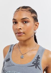 Grå tanktopp med grafisk design, sølvkjede med anheng, og små hoops-stikk øreringer. Jevn hudstruktur og flette.