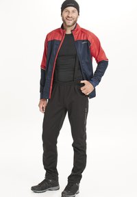 Veste de sport avec des sections rouge et marine, chemise noire en dessous, pantalon noir avec bretelles et bottes de randonnée grises, se tenant sur un fond blanc.