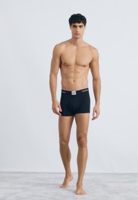 Boxeri din bumbac negru, cu un elastic moale care prezintă logo-ul mărcii, modelul stă desculț pe un covor deschis, pe un fundal simplu.
