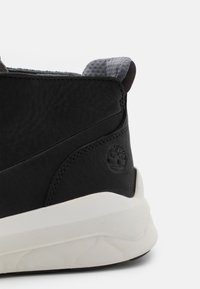 Baskets en cuir noir avec un col de cheville rembourré gris et une semelle en caoutchouc blanche. Présente un détail de logo sur le talon et un dessus texturé.