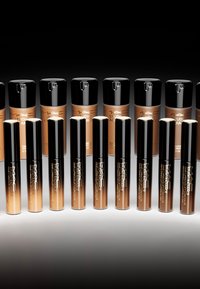 Botellas de base y corrector MAC Studio Radiance dispuestas en filas. Las botellas tienen tapas negras y varios tonos de marrón, con marcas en relieve.