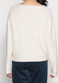 Takapaja henkilö, joka käyttää hulmuavaa pitkähihasta valkoista neuletta ja tummansiniset pöksyt, joissa on näkyvissä takatasku.