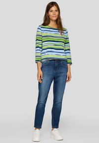 Frau steht und trägt ein langärmeliges Oberteil mit blauen, grünen und weißen Streifen, blaue Jeans und weiße Turnschuhe vor einem grauen Hintergrund.