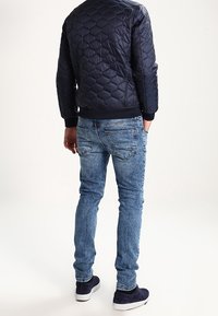 Marinblå kviltad bomberjacka med hexagonmönster och ribbad ärm, kombinerad med ljusblå smalpassande jeans och mörka sneakers. Perfekt finish.