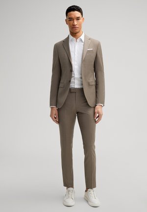 Mężczyzna stoi, ubrany w taupe garnitur, białą koszulę i białe trampki na jednolitym, jasnoszarym tle.