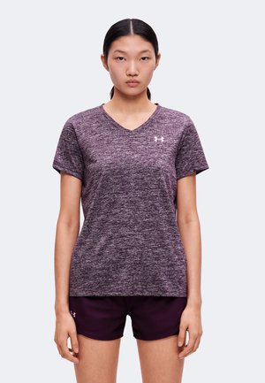 Jeune femme portant un t-shirt de sport texturé violet à col en V et un short violet foncé, debout devant un fond uni.