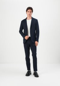 Fato azul-marinho ajustado com um casaco de um só botão, lapelas recortadas e calças combinando. Acompanhado de uma t-shirt branca texturizada e sapatos pretos.