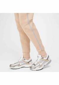Sneaker argento e bianchi con tomaia in rete, dettagli dorati e suola in gomma nera, abbinate a pantaloni jogger beige con striscia laterale grigia e tasche.