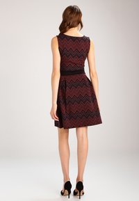 Robe sans manches avec un motif en zigzag noir et rouge, corsage ajusté, jupe évasée et une ceinture noire. Portée avec des chaussures à talons hauts noires.