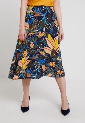 Vrouw draagt een knielange rok met een kleurrijk tropisch bloemenpatroon, gecombineerd met zwarte pumps met spitse neus en een mosterdkleurige top.