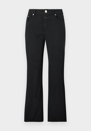 Stojaca žena nosi voľnú bielu košeľu so zapínaním na gombíky cez biely top, čierne nohavice s rovnými nohavicami a hnedé žabky, ruky v vreckách.