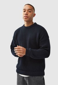 Navy knit sweater with a textured surface, ribbed cuffs, and waistband. Features a crew neckline and relaxed fit. Worn over a white shirt.
