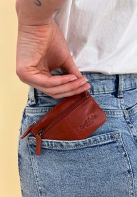 Červená kožená zipová taštička částečně umístěná v kapse ze světle praného denimu, s vyraženým logem a hladkým povrchem.