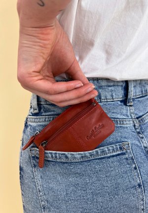 Červená kožená zipová taštička částečně umístěná v kapse ze světle praného denimu, s vyraženým logem a hladkým povrchem.